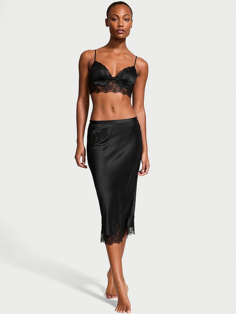 Modelka prezentuje zestaw satynowy - biustonosz braletka i spódnica midi. Produkt ma kolor czarny i pochodzi z kolekcji Victoria's Secret.
